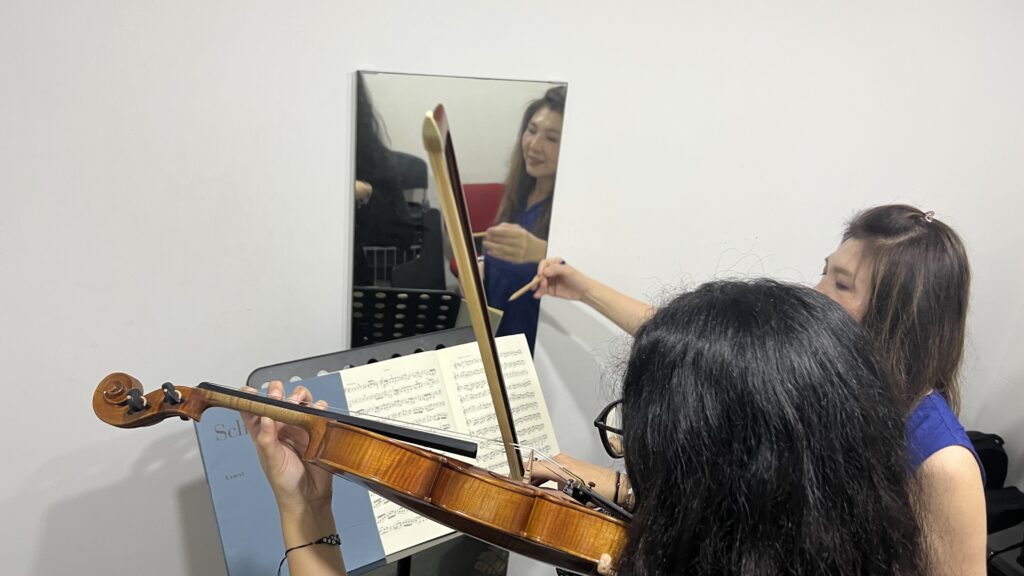 A violin student practising in front of the mirror with her teacher